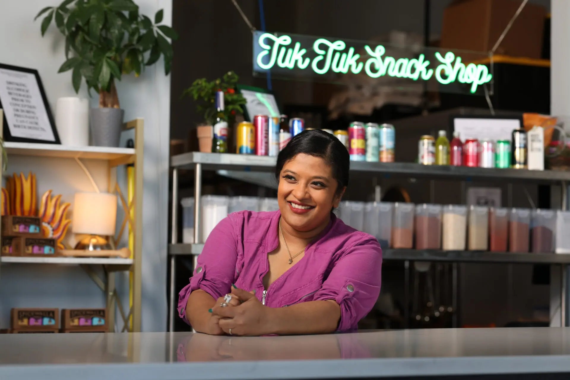 Chef Sam Fore in a pink shirt standing behind a counter with 'Tuk Tuk Snack Shop' neon sign in the background.